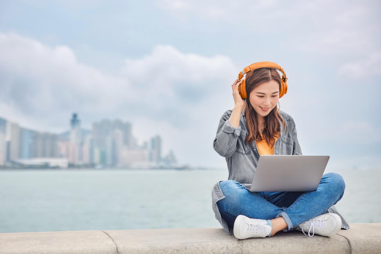 Young_woman_listening_to_headphones_on_her_laptop_sitting_on_a_ledge_by_the_promenade.jpg
