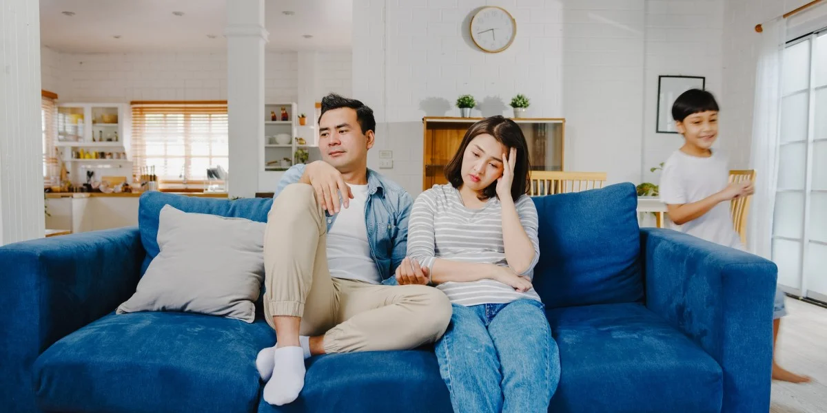 Cặp vợ chồng mệt mỏi ngồi trên sofa với đứa con chạy nhảy đằng sau